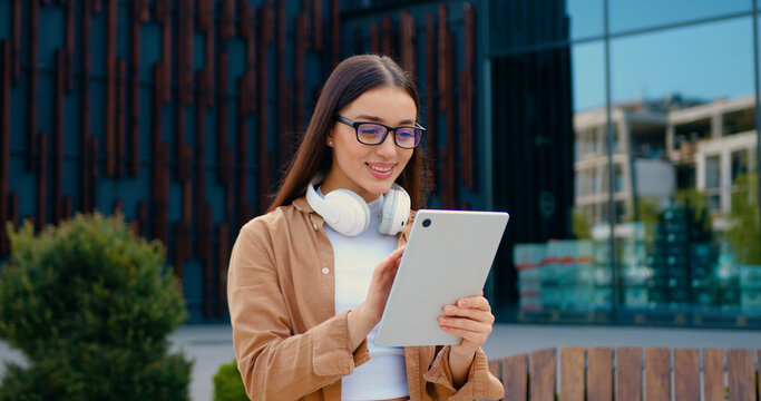 Attractive girl in glasses with headphones around neck and glasses, working on tablet on commercial building background, outdoors - Powered by Adobe