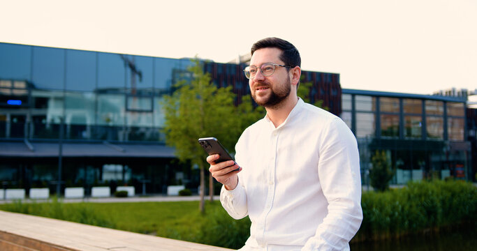 Serious businessman using on mobile phone standing on street near office building. Handsome male texting with business partner client, remote work