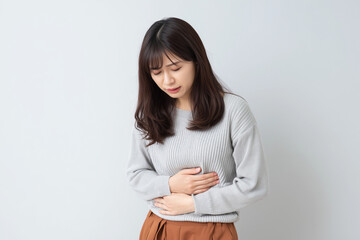 腹痛や胃痛で苦しんでいる表情でお腹を押さえているアジアの若い女性 / Young Asian woman holding her stomach with a pained expression due to stomach or abdominal pain