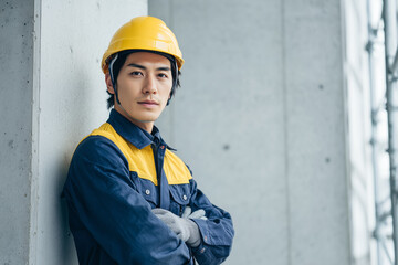 黄色いヘルメットと作業服を着用し、腕を組んでコンクリートの壁にもたれかかる若いアジア人作業員 / Young Asian construction worker in a yellow helmet and uniform, leaning against a concrete wall with arms crossed
