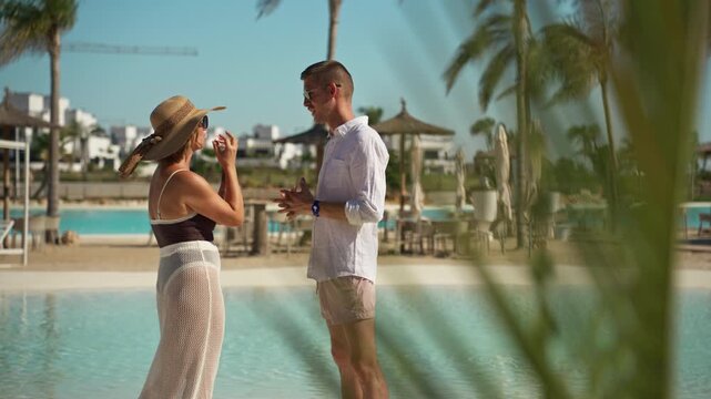 Young frustrated couple arguing and gesturing emotionally while having a relationship crisis on vacation, standing by a swimming pool at a luxury tropical resort with palm trees