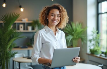 Smiling young woman holding laptop in modern office setting. She works indoors wearing casual outfit. Happy female pro uses computer at workplace. Inclusivity is visible.