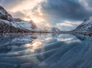 Aerial view of frozen lake in Lofoten, Norway, snow-covered mountains and low winter sun reflecting on the icy surface at sunset. Cold Arctic landscape, dramatic clouds. Winter scenery. Top drone view