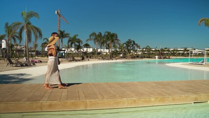 Young loving couple enjoy sunny day, walking hand in hand along wooden deck of luxury resort pool with clear blue water and palm trees on their summer vacation getaway