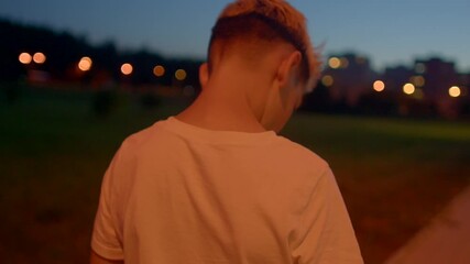 teen boy walking on park path under amber streetlights, back view shows white shirt and slow steps, city