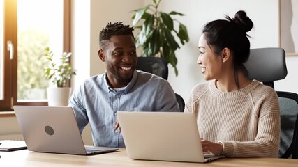 Happy diverse colleagues, an African man and an Asian woman, collaborating on laptops in a sunlit office. Positive teamwork and communication concept for business or corporate use. - Powered by Adobe