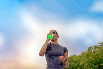 Young woman hydrating with a green drink against a sky background after exercising