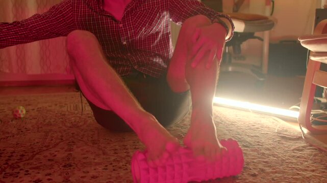 caucasian man seated massaging feet next to foam roller and ambient neon light, afterwork relief