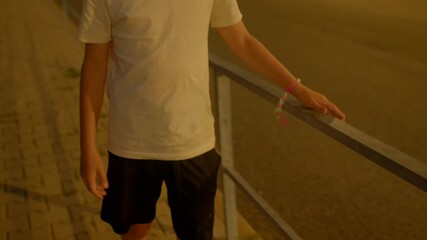 teen walking along railing at night, hand resting on metal bar with colorful bracelet, steady steps