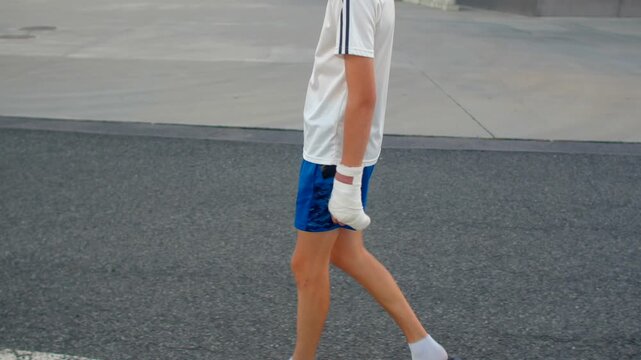 white athlete rehabilitating on asphalt road, blue shorts and white tee, wrist bandage and taped glove,