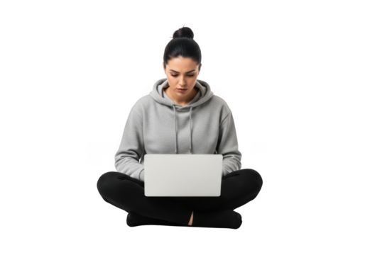 A focused young woman wearing a grey hooded sweatshirt and dark pants sits cross legged working on a laptop computer isolated on transparent background