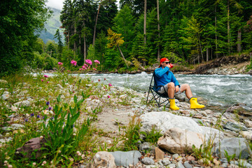 a man is sitting on the bank of a mountain river, drinking coffee
