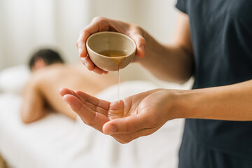 Horizontal intimate spa moment with warm massage oil being poured into the therapist’s hands as a client rests in the blurred background captured in natural soft lighting to express warmth, relaxation