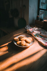 Warm sunset-lit wooden table with a dish of small freshly baked round buns, the kitchen fading into deep shadows,  the pastries glow in soft natural light, a nostalgic memory of rural home baking