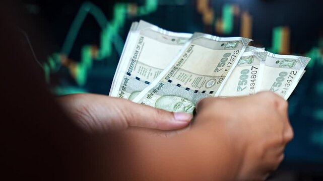 A hand counts Indian rupee notes against a blurred stock market screen, highlighting the connection between currency value and market movement