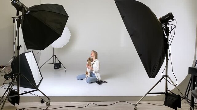 Blonde woman sits floor white cyclorama photo studio, taking selfie with professional camera and small, active, happy dog Jack Russell terrier Maya. professional photography equipment flashes video