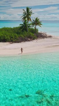 Aerial view of a young woman walking along a pristine white sand beach on a tropical island, surrounded by crystal-clear turquoise water and lush palm trees