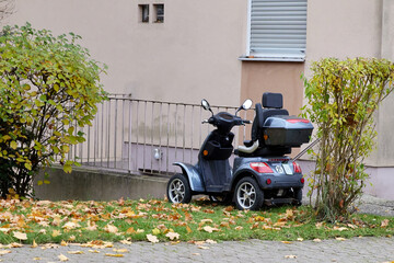 Electric mobility scooter parked outside a residential building surrounded by autumn leaves and bushes, representing accessibility and independence for elderly or disabled individuals. © beautiful-photo.de