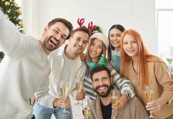 Group of young happy friends standing in living room at home celebrating Christmas looking cheerful at camera making selfie holding glasses of champagne in hands. New Year celebration concept.