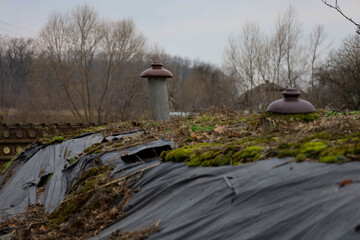 This photograph shows a dilapidated earth shelter or old structure covered in black plastic sheeting, moss, and dry leaves, featuring two vent pipes topped with brown caps.