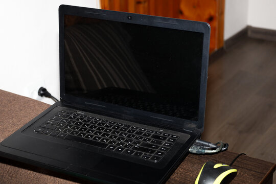 This shot showcases a modern black laptop with a blank screen, resting on a workspace or desk surface in a home setting. A black and yellow mouse and connected cables are visible nearby, creating a pr