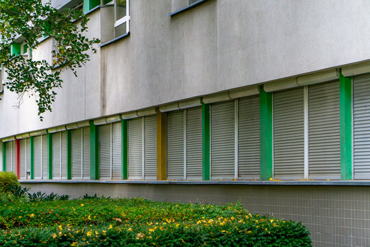 Building facade with closed shutters, colorful vertical panels and surrounding greenery creating a clean, structured urban architectural composition suitable for modern visual contexts