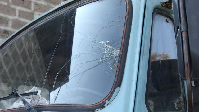 Close-up shot of a heavily cracked and damaged windshield on an abandoned, rusty, vintage blue truck. The broken glass shows severe impact damage, symbolizing decay, accidents, and neglect.