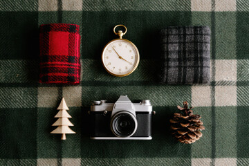 Vintage camera with pocket watch, pine cone, wooden tree, and plaid fabric on green checkered background, evoking nostalgic holiday warmth