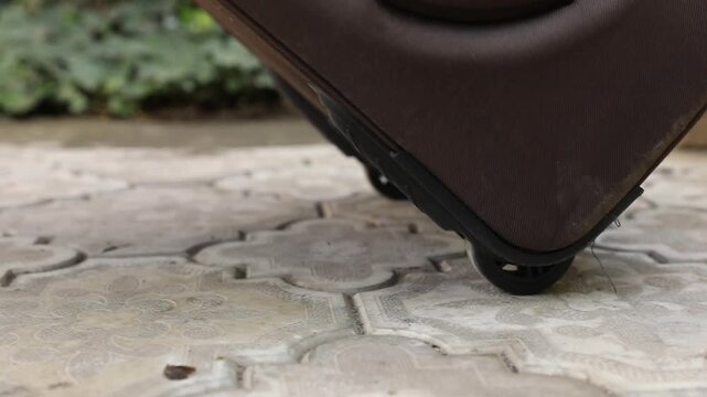 Extreme close-up of a brown luggage wheel rolling slowly across decorative, sunlit outdoor paving. This shot represents the themes of travel, journey, and movement.