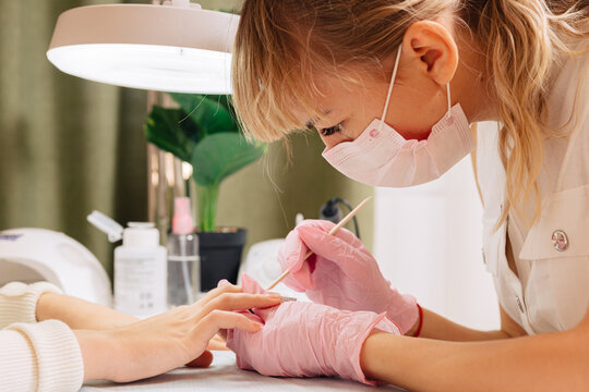 Close-up of a masked manicurist in pink gloves applying nail polish to a client's fingernails under a bright professional lamp in a beauty salon.