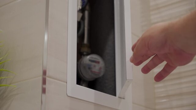 Hand opening a small white hinged cabinet door built into a tiled wall, revealing water meters inside. The door swings open and then closes. A slight squeaking and rubbing sound is heard from hinges.