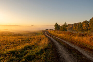 Fototapeta premium A picturesque view of a road through a field against the sky during sunrise. A landscape against the sky on a foggy morning.