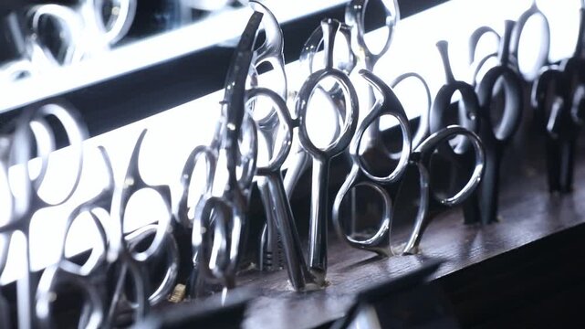 Professional barber scissors and shears stand organized in a backlit display. Dramatic lighting highlights the shiny metal tools used for precision haircutting in a modern salon or barbershop.