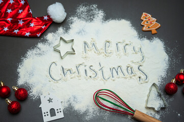 Flour surface with a handwritten Merry Christmas message, festive ornaments, cookie cutters, and a Santa hat creates a warm seasonal mood. Ideal for holiday advertising, baking themes, and designs.