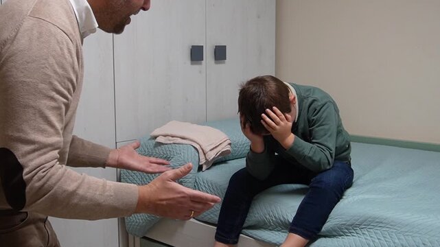 A father stands angrily scolding his young son, who sits on the bed covering his face with his hands, in a tense family moment inside a bedroom.