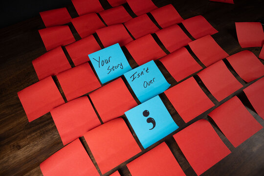 Text and single semi colon on blue sticky notes in a grid pattern of red sticky notes on top of dark wood surface.  Your story isn't over.