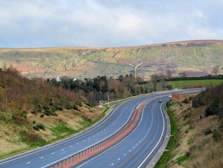An elevated wide shot captures a modern, dual-carriageway highway curving through a rolling, semi-rural landscape under a pale, overcast sky. The road is flanked by grassy slopes and sparse trees
