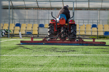 Tractor on sports field Maintaining artificial turf - A red utility tractor with a rake attachment maintains an artificial turf football field