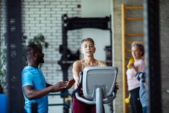 Adult trainer coaching mature woman on elliptical in gym, focused