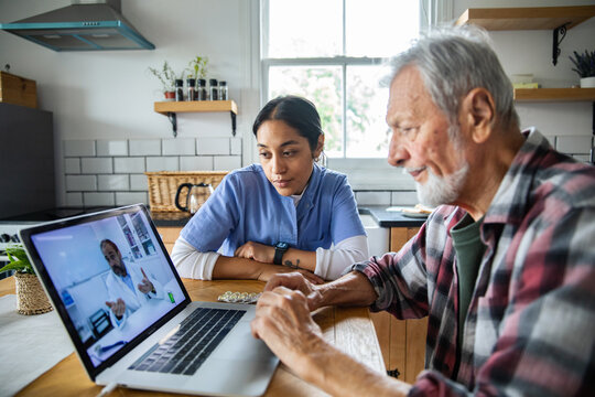 Senior man and adult caregiver focused on telehealth in home kitchen