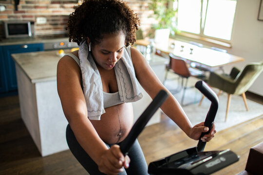 Pregnant adult woman exercising on elliptical at home, focused