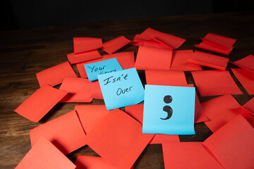 Text and single semi colon on blue sticky notes in a grid pattern of red sticky notes on top of dark wood surface.  Your story isn't over.