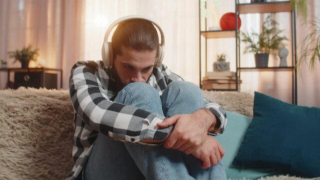 Young man sits at home wearing headphones, listening to emotional tune music, fighting back tears. Guy on sofa removes earphones, releasing heavy sigh over painful breakup or separation. Broken heart
