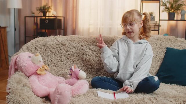 Teenager girl places toys plush figures in front at home sofa, seriously reciting repeats simple math arithmetic. Child kid occasionally giggles laughs amused by own mock authority, silly teacher role