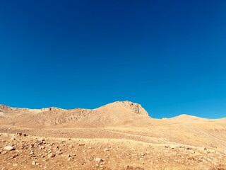 Arid, desolate mountain ridge or hill with light brown soil and rocky terrain under a bright, clear blue sky. Travel, drought, ecology, and summer or autumn desert landscape concept.