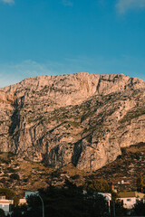 Imposing, sun-drenched, textured rock mountain towering over low-rise residential buildings and dark green trees in the foreground.