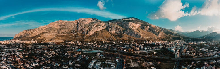 Naklejka premium Panoramic aerial landscape of a sprawling coastal town with dense housing, nestled directly below a massive, rugged mountain range under a bright sky.