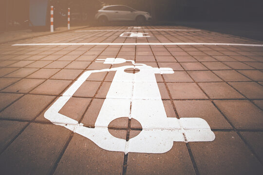Electric vehicle parking symbol on paved charging space with warm sunlight and parked car in the background.