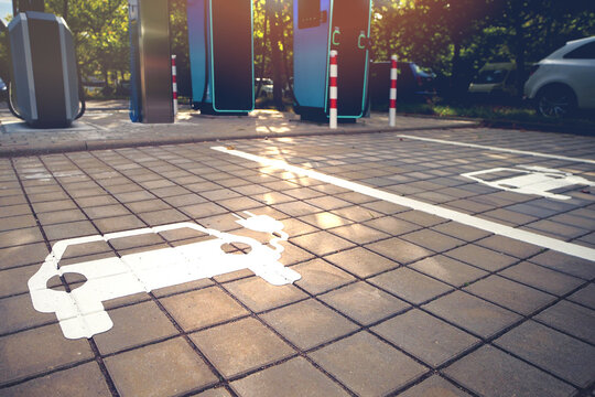 Electric vehicle parking symbol on sunlit charging area with strong light reflections and modern chargers in the background.