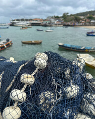 Close-up of a blue fishing net with white floats in the foreground, overlooking a small harbor on a cloudy day. 